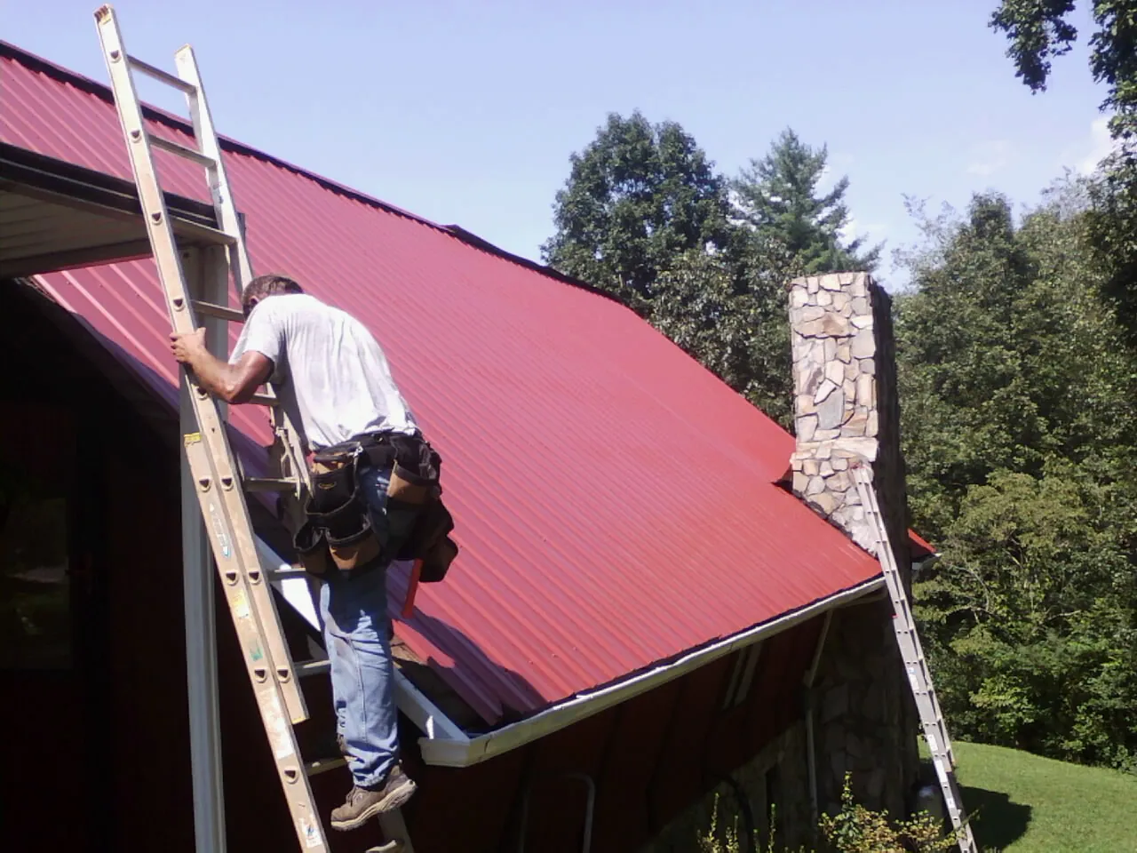 Roofer on a ladder beside a red metal roof and stone chimney — Soffit & Fascia Repair by Roofing Brothers in Mcconnelsville