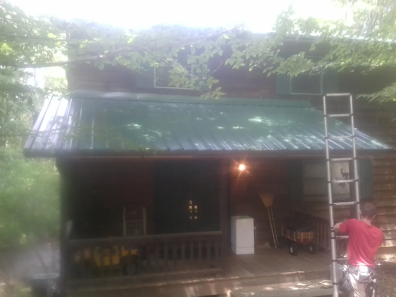 Worker on a ladder at the green porch roof of a wooded cabin in Mcconnelsville